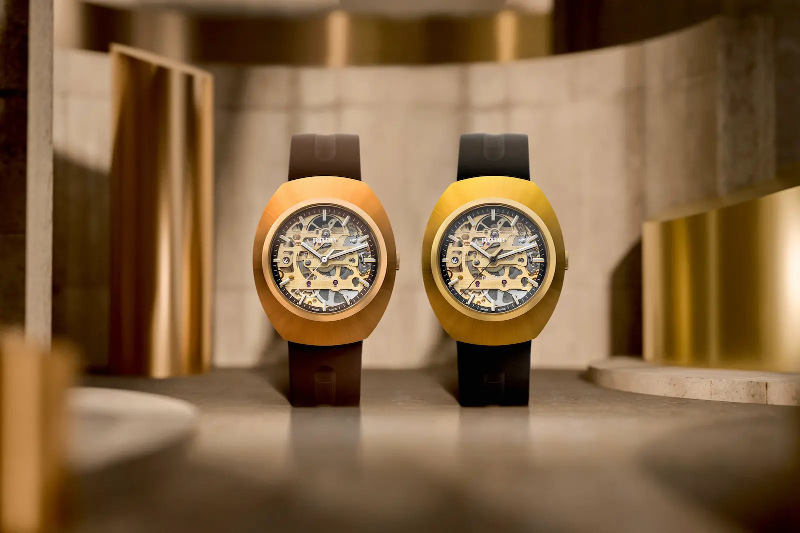 I due Rado DiaStar Original Skeleton, color oro rosa con cinturino marrone e color oro giallo con cinturino nero, su un fondale color pietra con pannelli dorati